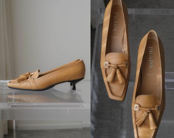 Puede incluir: Un par de mocasines de cuero color canela con acabado texturizado, que presentan un tacón pequeño, punta cuadrada y un detalle de borlas decorativas en la parte delantera. La plantilla interior lleva el logo de Prada en blanco.