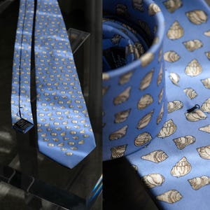 May include: A blue necktie featuring a repeating pattern of beige seashells. The tie is made of a smooth fabric and is displayed on a glass surface. The design is visible on both the front and the rolled-up portion of the tie.