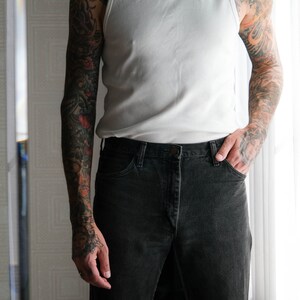 May include: A man wearing a white tank top and black jeans. He has tattoos on both arms.