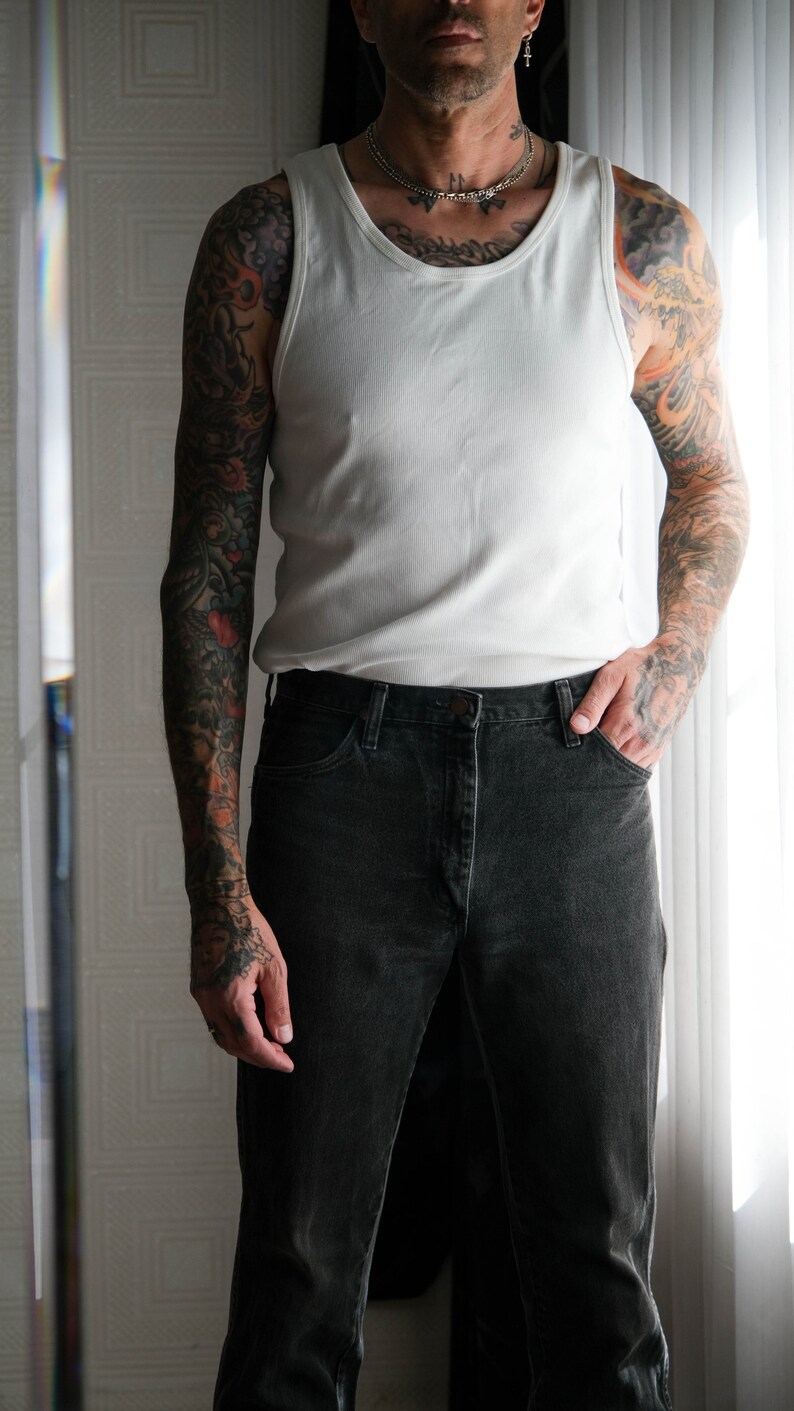 May include: A man wearing a white tank top and black jeans. He has tattoos on both arms.
