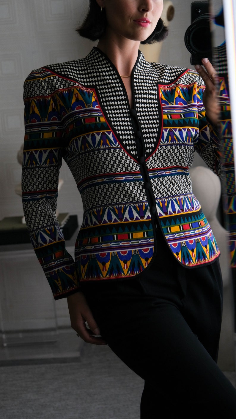 May include: A black and white patterned blazer with a colorful geometric design. The blazer has a V-neck and a button closure. The model is wearing black pants.
