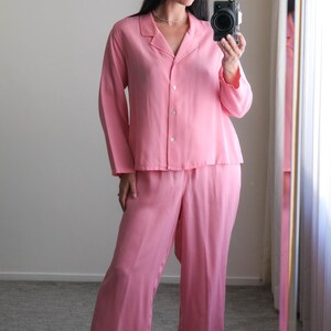 May include: A light pink pajama set, including a button-down shirt and matching pants. The outfit is made of a soft, silky material. The model is wearing pink heeled sandals.