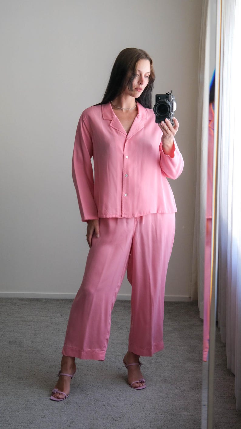 May include: A light pink pajama set, including a button-down shirt and matching pants. The outfit is made of a soft, silky material. The model is wearing pink heeled sandals.