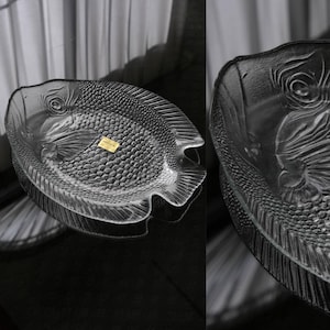 May include: A clear glass serving platter shaped like a fish, featuring a textured scale pattern on the body, defined fins, and a prominent eye. A small rectangular gold-colored sticker is adhered to the center of the glass surface.
