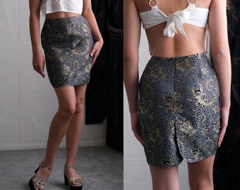 May include: A woman is wearing a short, blue and gold brocade skirt with a white top. The skirt has a floral pattern and is fitted.