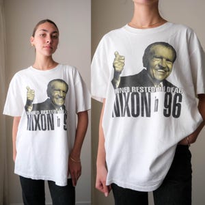 May include: A white short-sleeve t-shirt featuring a black and white graphic of Richard Nixon giving a thumbs up. The text printed on the front reads TANNED RESTED AND DEAD NIXON in 96 in bold black lettering across the chest area of the garment.