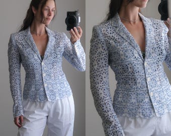 Puede incluir: Una americana de color azul claro y blanco con un intrincado patrón floral de encaje calado en toda la prenda. Cuenta con mangas largas, cuello de solapa y cierre frontal con tres botones blancos. El dobladillo presenta un borde festoneado.