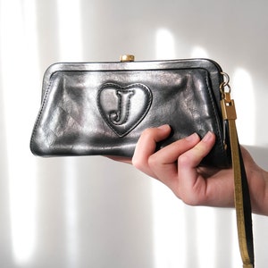 May include: A black leather clutch with a gold clasp and a heart-shaped detail with a gold "J" in the center. The clutch has a gold chain strap.