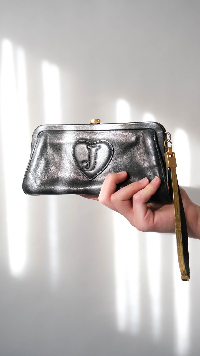 May include: A black leather clutch with a gold clasp and a heart-shaped detail with a gold "J" in the center. The clutch has a gold chain strap.