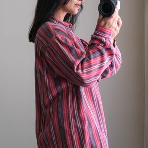 May include: A long-sleeved shirt with vertical stripes in shades of red, pink, and purple. The shirt has a button-down collar and cuffs. The person is holding a black camera.