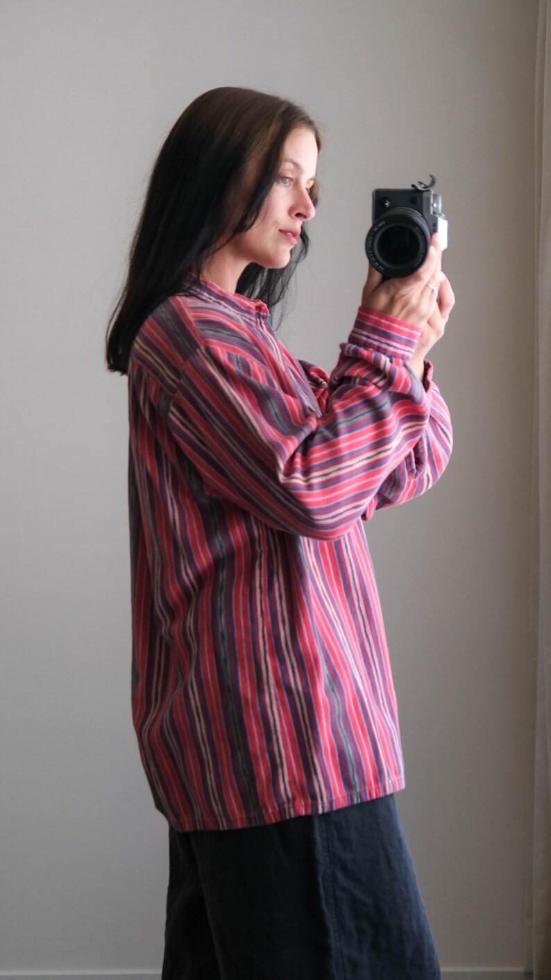 May include: A long-sleeved shirt with vertical stripes in shades of red, pink, and purple. The shirt has a button-down collar and cuffs. The person is holding a black camera.