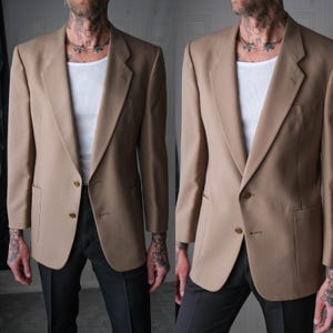 Könnte beinhalten: Ein beigefarbener Blazer mit zwei Knöpfen, einem fallenden Revers und zwei Vordertaschen. Der Blazer wird mit einem weißen Unterhemd und einer schwarzen Hose getragen. Die Jacke hat eine klassische, maßgeschneiderte Passform.