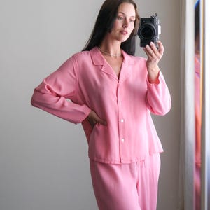 May include: A light pink pajama set, including a button-down shirt and matching pants. The long-sleeved shirt has a collar and buttons down the front. The pants are loose-fitting. The outfit is made of a soft, flowing fabric.
