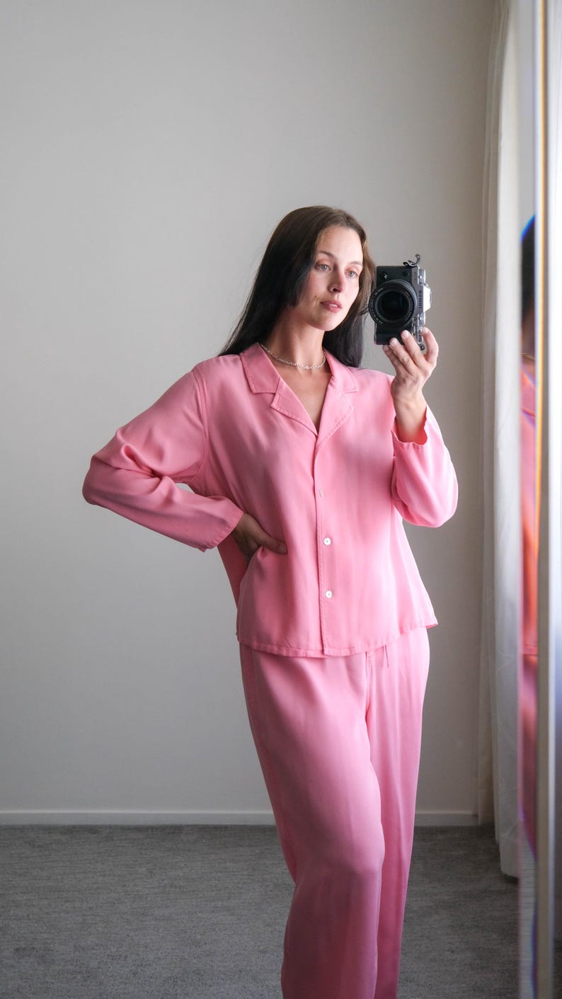 May include: A light pink pajama set, including a button-down shirt and matching pants. The long-sleeved shirt has a collar and buttons down the front. The pants are loose-fitting. The outfit is made of a soft, flowing fabric.