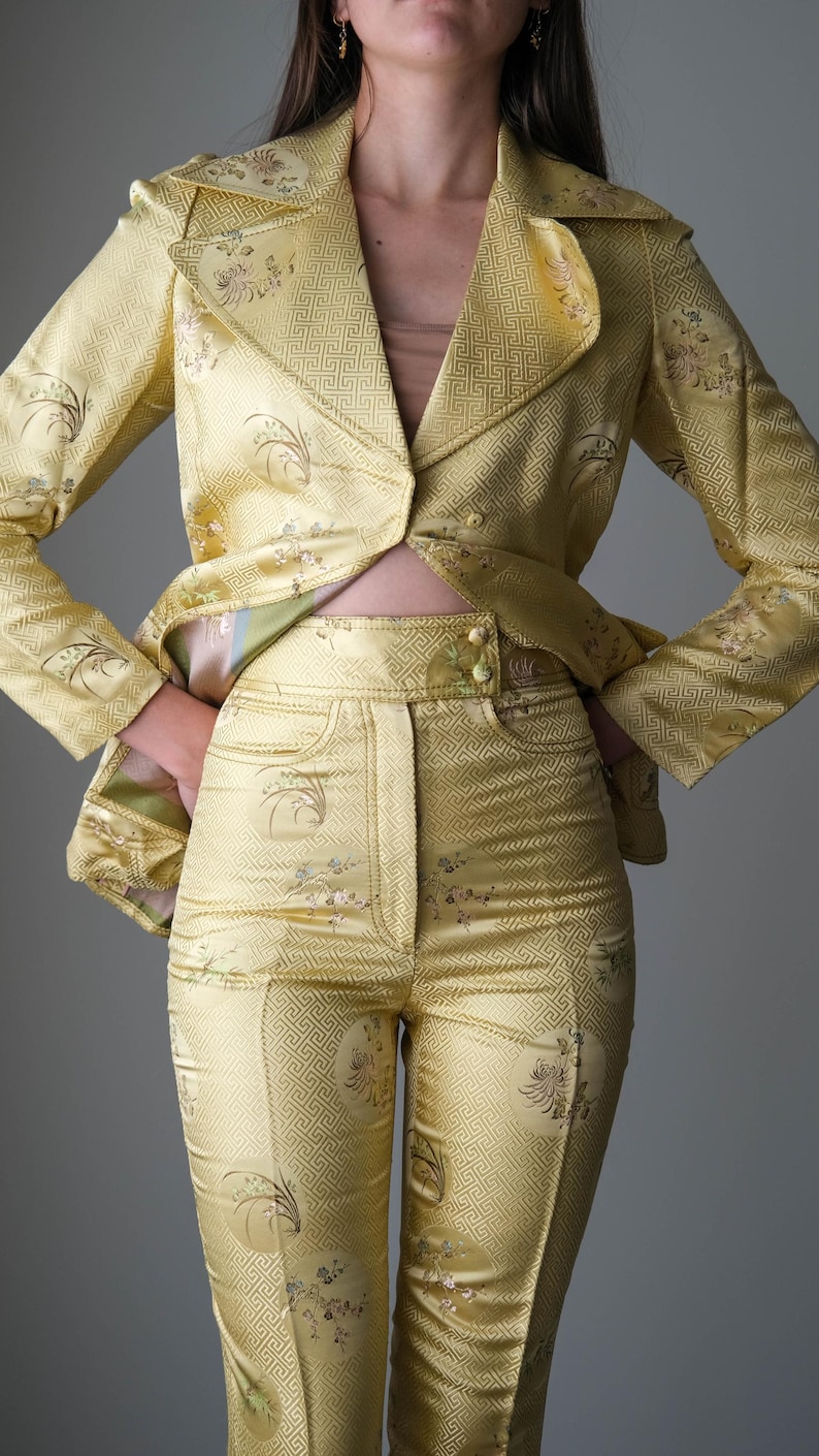 May include: A two-piece yellow satin suit with a floral and geometric pattern. The suit includes a blazer with a notched collar and matching high-waisted pants. The fabric has a subtle sheen, and the suit appears to be vintage.
