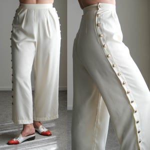 May include: Cream-colored wide-leg pants with a row of gold-colored buttons running down the side seams. The pants have a high waist and pleats at the front. The fabric appears to be a lightweight material.