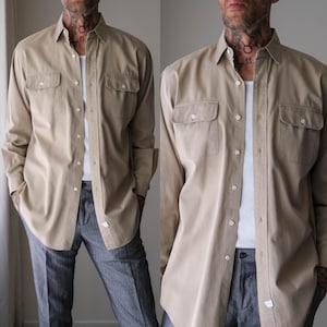 May include: A tan button-down shirt with two chest pockets. The shirt is worn over a white undershirt and paired with gray trousers. The shirt has long sleeves and a classic collar.