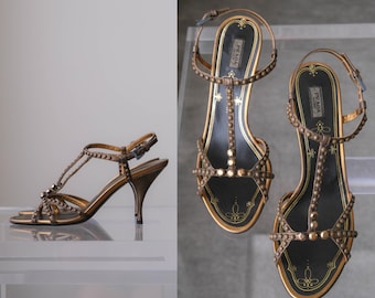 Puede incluir: Un par de sandalias Prada en tono bronce con diseño de correa en T, adornos de tachuelas metálicas, correa de tobillo ajustable con hebilla y tacón medio. Las plantillas negras están decoradas con dibujos lineales dorados.