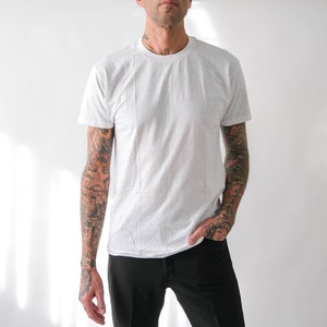 May include: A white t-shirt with a crew neck and short sleeves. The person wearing the shirt has tattoos on their arms.