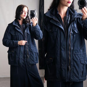 May include: A navy blue military-style jacket with multiple pockets, a zip-up front, and a hood. The jacket has a drawstring waist and button details. The model is wearing a black dress underneath. The jacket appears to be made of a durable, possibly water-resistant material.