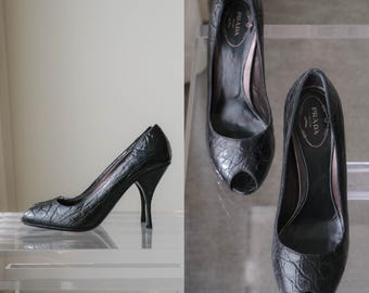 Puede incluir: Un par de zapatos de tacón alto negros con un acabado texturizado de efecto cocodrilo y diseño peep-toe. Las plantillas interiores presentan el logotipo de Prada Milano en letras doradas. Los zapatos tienen un tacón curvo y silueta elegante.