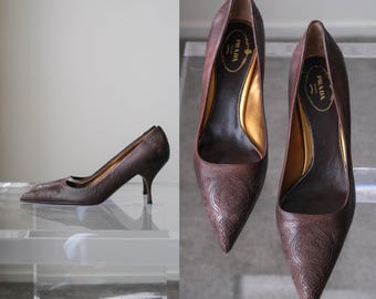 Puede incluir: Un par de zapatos de tacón de cuero marrón oscuro con punta afilada y un patrón texturizado en relieve en el exterior. El interior presenta un forro dorado y una plantilla negra con el logotipo de Prada Milano en letras doradas.
