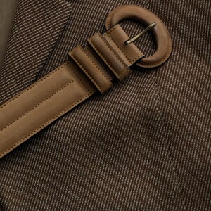 May include: Close-up of a brown blazer with a matching leather belt. The belt has a large, rounded buckle and is secured with a series of rectangular loops. The blazer is made of a textured fabric with a subtle striped pattern.