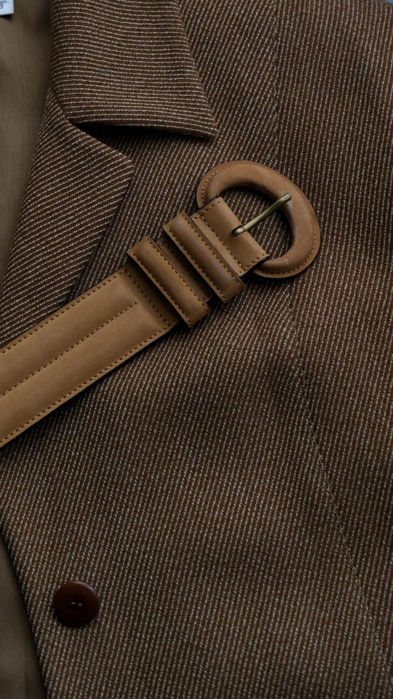May include: Close-up of a brown blazer with a matching leather belt. The belt has a large, rounded buckle and is secured with a series of rectangular loops. The blazer is made of a textured fabric with a subtle striped pattern.