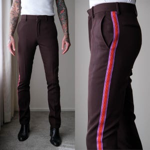 May include: Dark brown trousers with a vertical stripe in shades of red and purple down the side. The pants have a classic fit and are shown with black boots. The stripe adds a pop of color to the otherwise solid-colored pants.