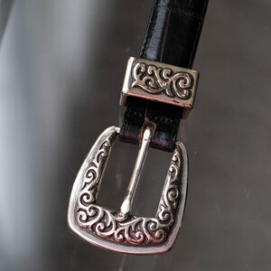 May include: A black leather belt with a silver buckle and matching belt loop hardware. The buckle and loop feature an ornate, swirling design. The belt is displayed against a reflective surface.