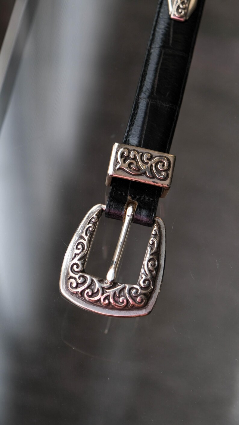 May include: A black leather belt with a silver buckle and matching belt loop hardware. The buckle and loop feature an ornate, swirling design. The belt is displayed against a reflective surface.