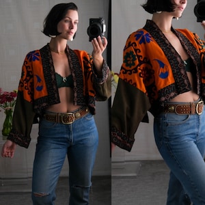 May include: A cropped, orange, green, and blue embroidered jacket with a brown collar and sleeves. The jacket is worn over a black bra and blue jeans with a brown belt.