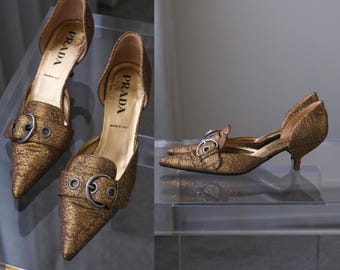 Puede incluir: Un par de zapatos de tacón de punta en color bronce con acabado texturizado, que presentan un diseño con aberturas laterales y una hebilla decorativa en el empeine. La plantilla interior dorada muestra claramente la marca Prada y Made in Italy.
