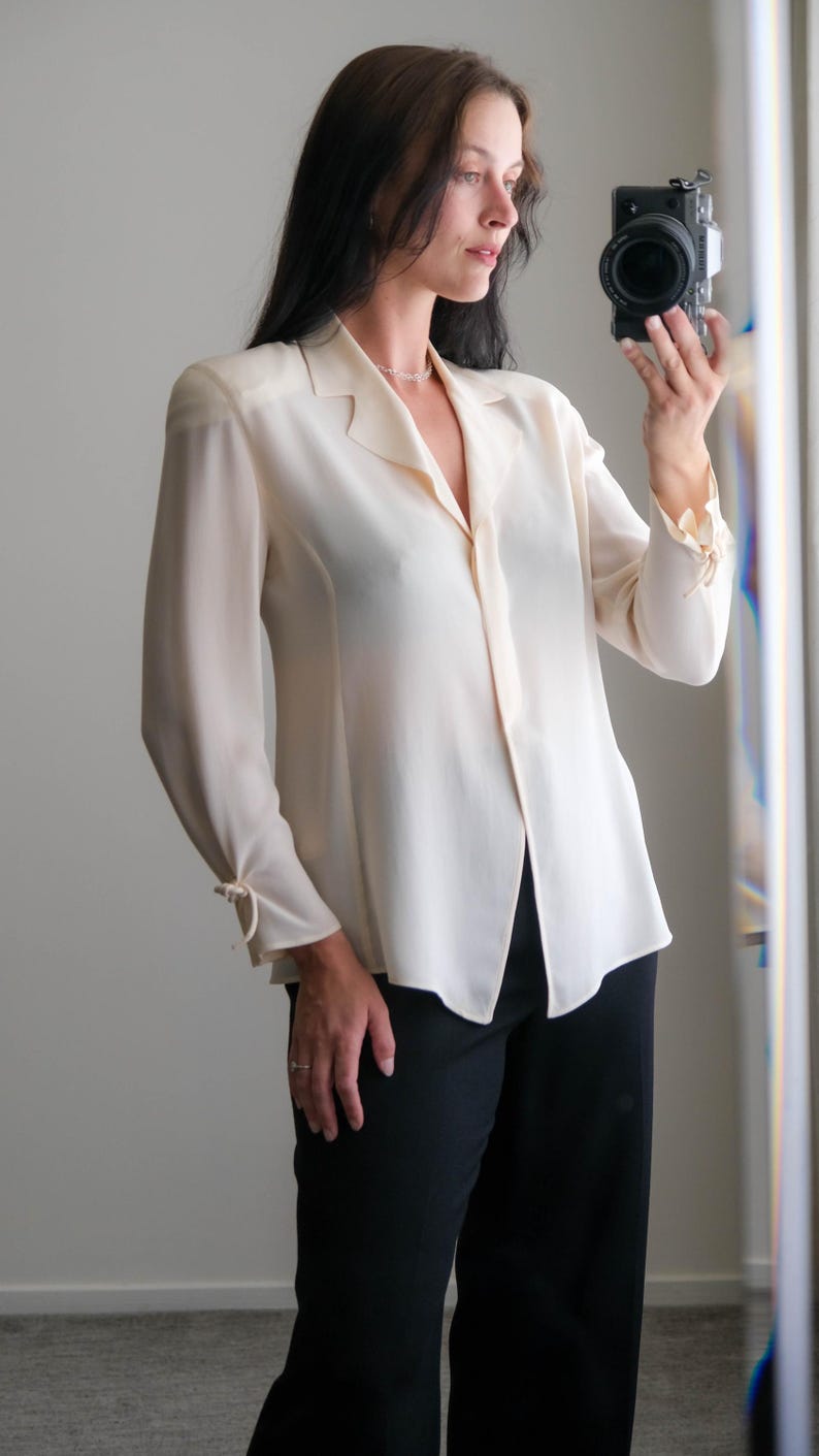 Peut inclure: Un chemisier cr&egrave;me &agrave; manches longues et col. Le chemisier a une fermeture boutonn&eacute;e sur le devant et est associ&eacute; &agrave; un pantalon noir. Les manches sont fronc&eacute;es aux poignets avec un d&eacute;tail de n&oelig;ud. La tenue est simple et &eacute;l&eacute;gante.