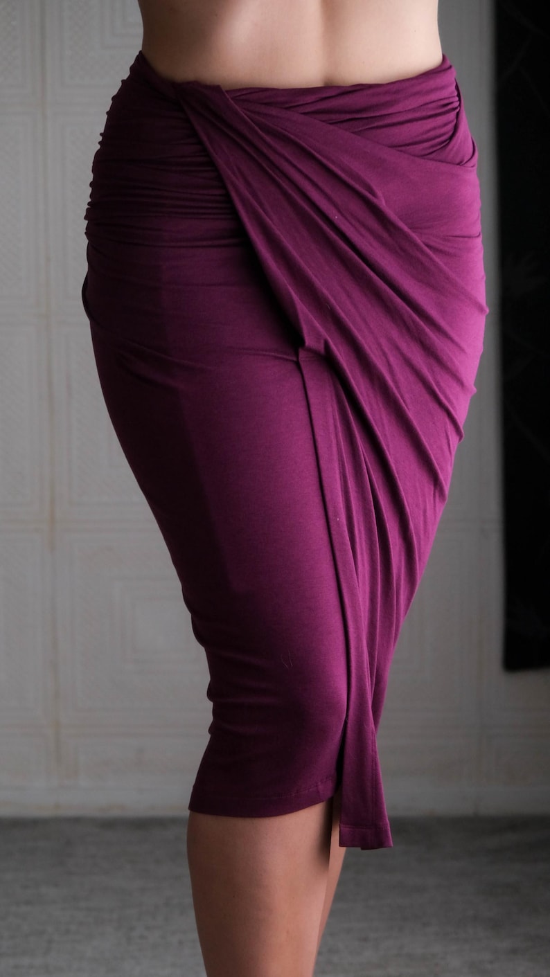 May include: A draped, midi-length skirt in a deep plum color. The fabric is gathered and wrapped, creating a unique asymmetrical design. The skirt appears to be made of a soft, flowing material.