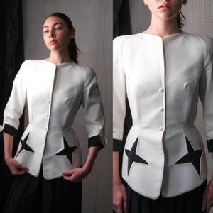 May include: A white jacket with black star cutouts and black cuffs. The jacket has a button-up front and three-quarter sleeves. The jacket is tailored with a fitted waist and structured shoulders.