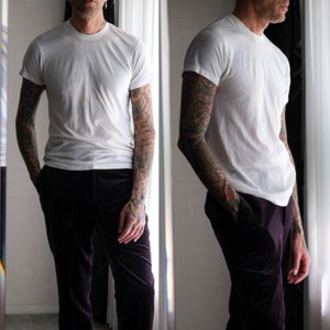 May include: A white t-shirt with short sleeves and a crew neck. The person is wearing dark purple trousers.