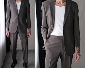 May include: A two-piece brown suit with subtle pinstripes. The suit includes a single-breasted blazer and matching trousers. The model is wearing a white undershirt and black boots. The suit is tailored for a classic, professional look.