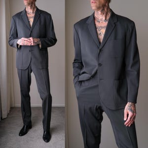 Könnte beinhalten: Ein anthrazitfarbener Anzug, bestehend aus einem Drei-Knopf-Blazer mit zwei Vordertaschen und einer passenden Hose. Der Anzug ist aus einem glatten Stoff gefertigt und wird mit schwarzen Anzugschuhen getragen. Der Gesamtstil ist klassisch und professionell.