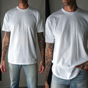 Może przedstawiać: Biały t-shirt z okrągłym dekoltem. Koszulka z krótkim rękawem wykonana jest z miękkiego materiału. Modelka ma na sobie jasnoniebieskie dżinsy. Model ma tatuaże na obu ramionach.