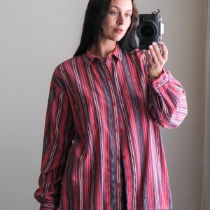 May include: A long-sleeved button-down shirt with vertical stripes in shades of red, pink, and purple. The shirt has a classic collar and a single chest pocket. The model is wearing dark blue pants.