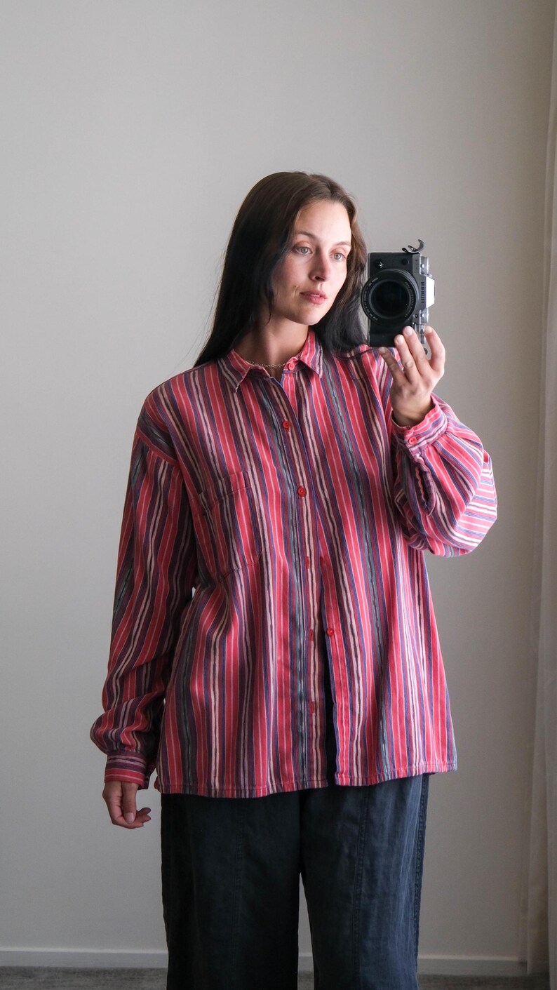 May include: A long-sleeved button-down shirt with vertical stripes in shades of red, pink, and purple. The shirt has a classic collar and a single chest pocket. The model is wearing dark blue pants.