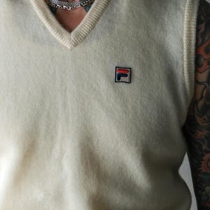 Peut inclure: Un gilet de pull cr&egrave;me &agrave; col en V avec un petit &eacute;cusson logo sur la poitrine. Le gilet a un col et des emmanchures c&ocirc;tel&eacute;s. Le logo est un carr&eacute; avec les lettres "F" et "L" en rouge, blanc et bleu.
