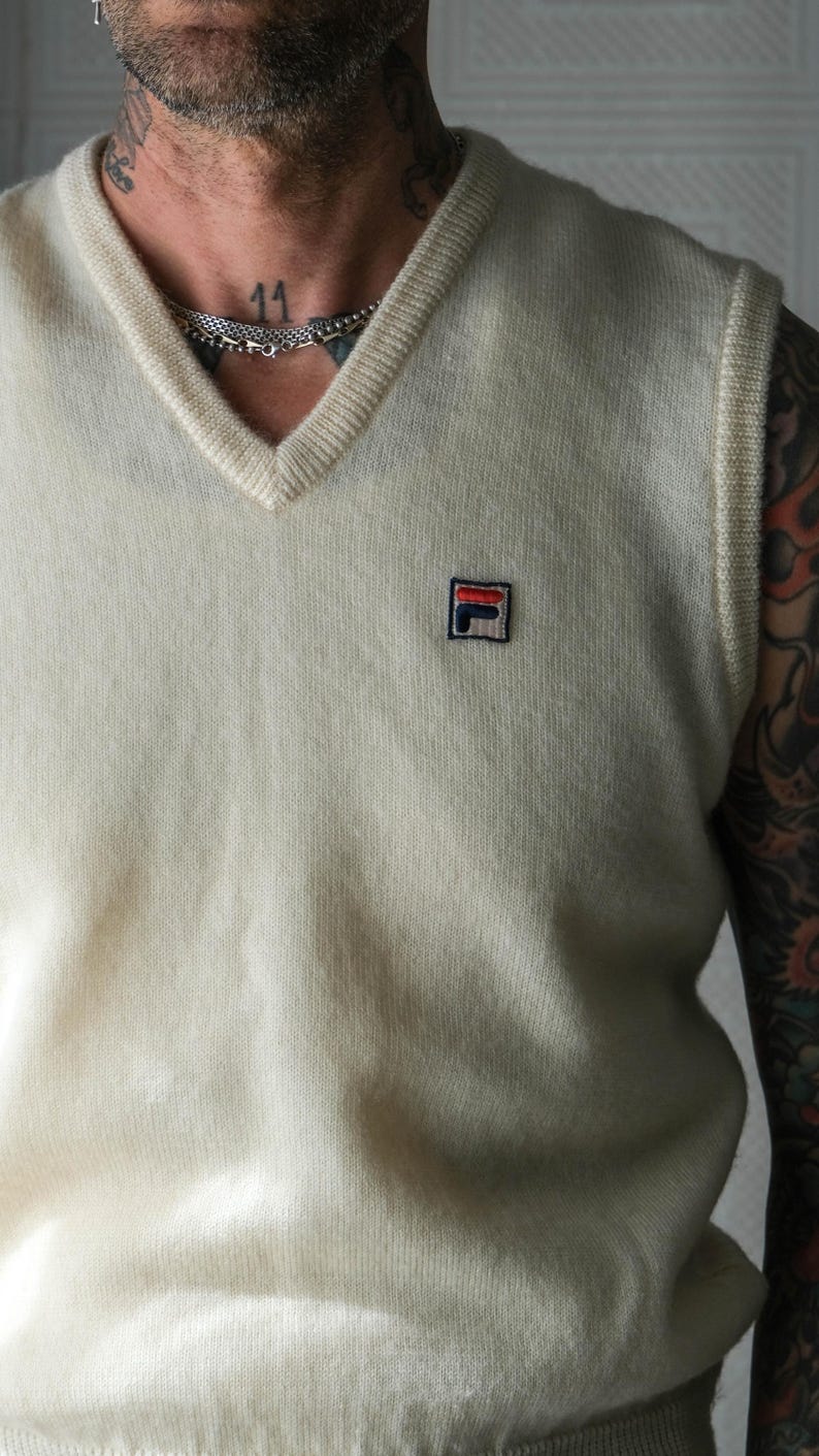 Peut inclure: Un gilet de pull cr&egrave;me &agrave; col en V avec un petit &eacute;cusson logo sur la poitrine. Le gilet a un col et des emmanchures c&ocirc;tel&eacute;s. Le logo est un carr&eacute; avec les lettres "F" et "L" en rouge, blanc et bleu.
