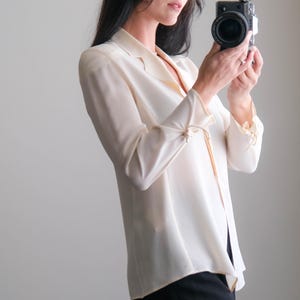 Peut inclure: Une blouse cr&egrave;me boutonn&eacute;e &agrave; manches longues et col chemise. La blouse est associ&eacute;e &agrave; un pantalon noir. Un appareil photo noir est tenu devant la personne.