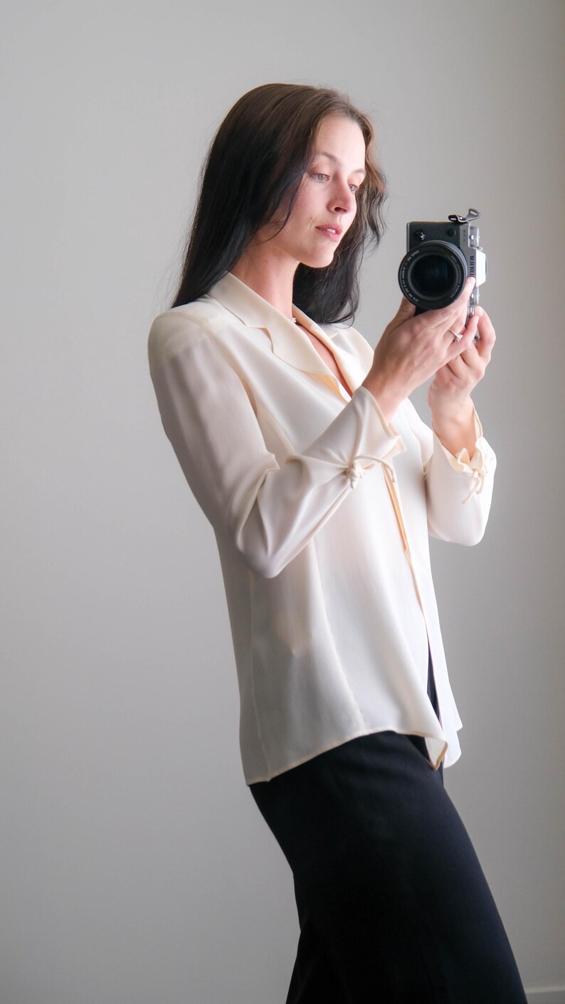 Peut inclure: Une blouse cr&egrave;me boutonn&eacute;e &agrave; manches longues et col chemise. La blouse est associ&eacute;e &agrave; un pantalon noir. Un appareil photo noir est tenu devant la personne.