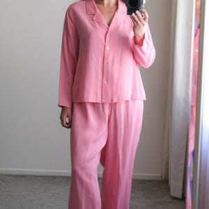 May include: A light pink pajama set, featuring a button-down top with a collar and matching wide-leg pants. The outfit is paired with light pink high-heeled sandals. The person is holding a camera.