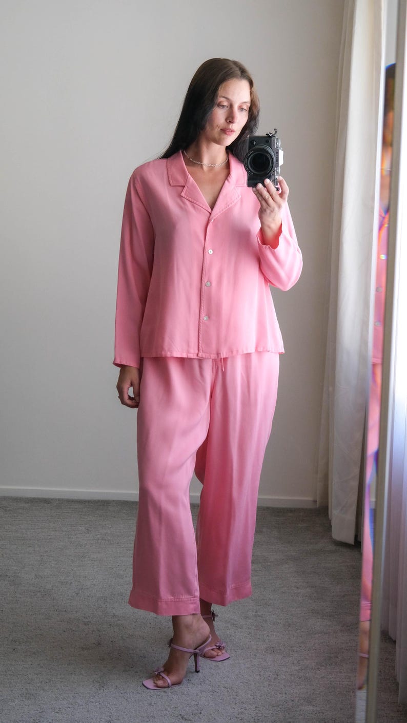 May include: A light pink pajama set, featuring a button-down top with a collar and matching wide-leg pants. The outfit is paired with light pink high-heeled sandals. The person is holding a camera.