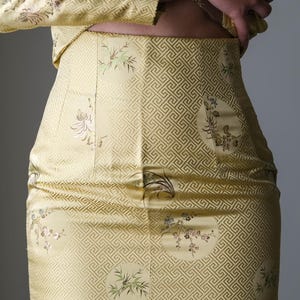 May include: A two-piece outfit in a pale yellow hue, featuring a skirt and a jacket. The fabric is adorned with a geometric pattern and floral embroidery. The jacket's sleeves are visible, and the skirt is fitted.
