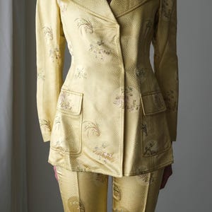 May include: A yellow suit with a floral and geometric pattern. The suit includes a double-breasted blazer with two front pockets and matching pants. The fabric has a subtle sheen, and the suit appears to be vintage.